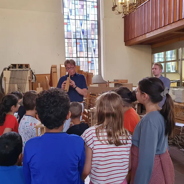 Kinder in der Kirche beobachten einen Mann, der eine hölzerne Orgelpfeife spielt.