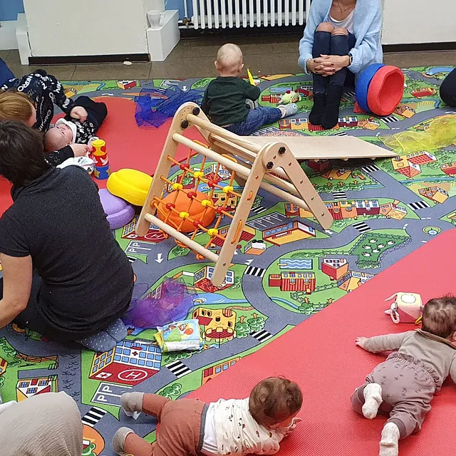 Babys und Eltern in einer Krabbelgruppe mit Klettergerüst und Spielmatten.