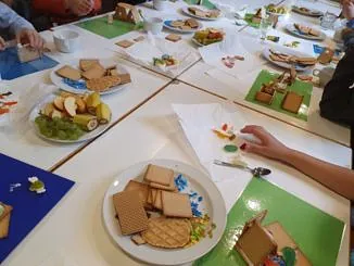 Kinder an einer langen Tafel mit Tellern voller Kekse, Obst und Süßigkeiten für einen Bastelworkshop.