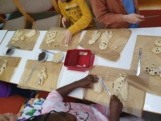 Kinder sitzen am Tisch und formen sowie verzieren Weckmänner aus Teig mit Rosinen.