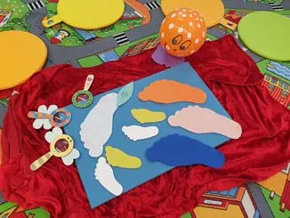 Bunte Papier-Fußabdrücke auf einer blauen Tafel, die auf einem roten Tuch über einer Spielmatte liegt.