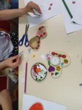 Kinder am Tisch beim Dekorieren von herzförmigen Holzkästchen und Kerzenhaltern mit kleinen bunten Steinen und Ausschnitten.
