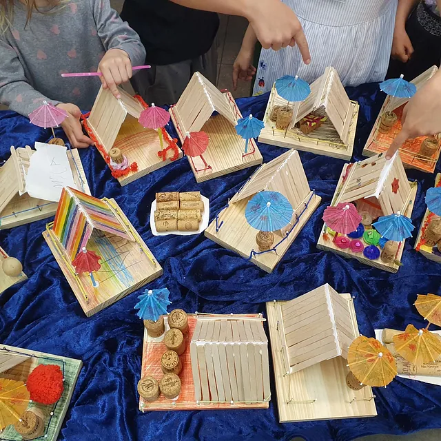 Kinder zeigen selbstgebastelte Holzhütten mit bunten Papierschirmen.
