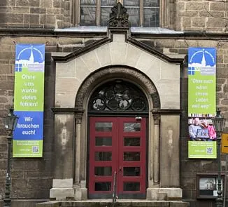 Der Steineingang der Sophienkirche mit roten Türen, flankiert von zwei großen, bunten vertikalen Bannern, die auf die Angebote der Gemeinde aufmerksam machen.
