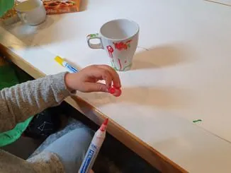 Ein Kind bemalt eine weiße Porzellantasse mit einem roten Stift während eines Gemeindeworkshops.