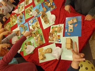 Kinder um einen Tisch mit roter Decke, die ihre selbstgebastelten Lebkuchenhäuser zeigen.